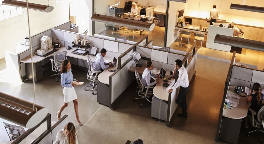 Elevated view of a busy, modern office with cubicles coloured grey and a yellow-lit area in the background.