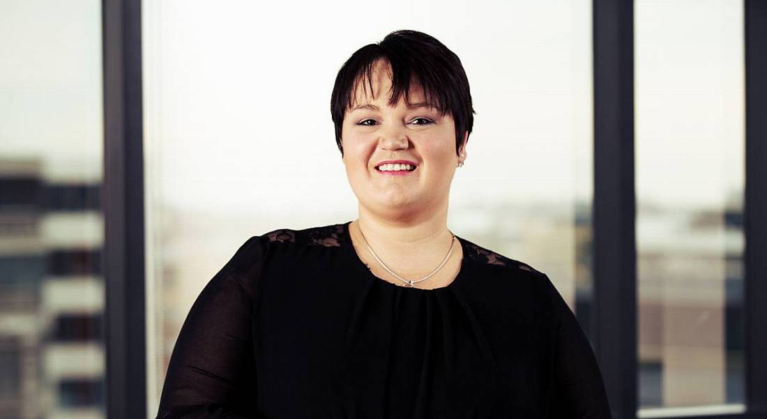 A woman in black business dress with cropped chestnut hair smiles at the camera.