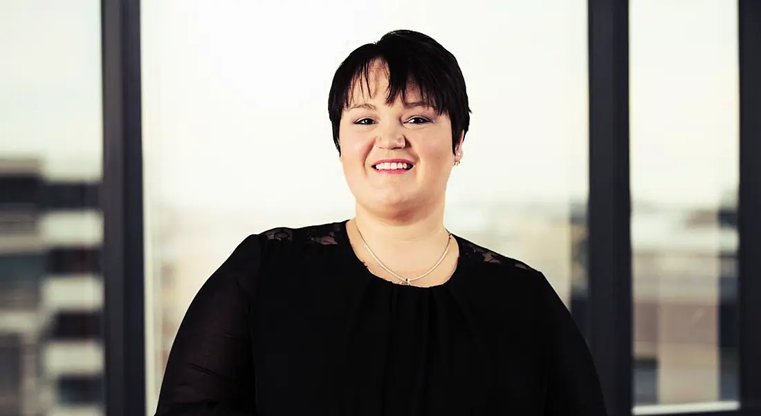 A woman in black business dress with cropped chestnut hair smiles at the camera.