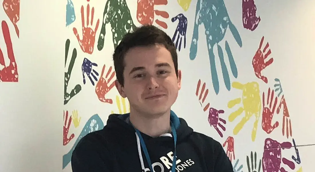 A young man in a navy hoodie smiling at the camera with multicoloured handprints on mural in the background.