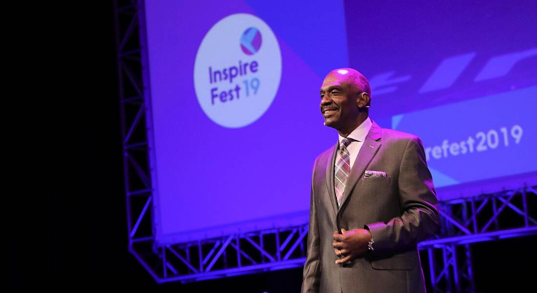 A man in a slate grey suit grinning widely at a crowd at the Inspirefest 2019 conference.