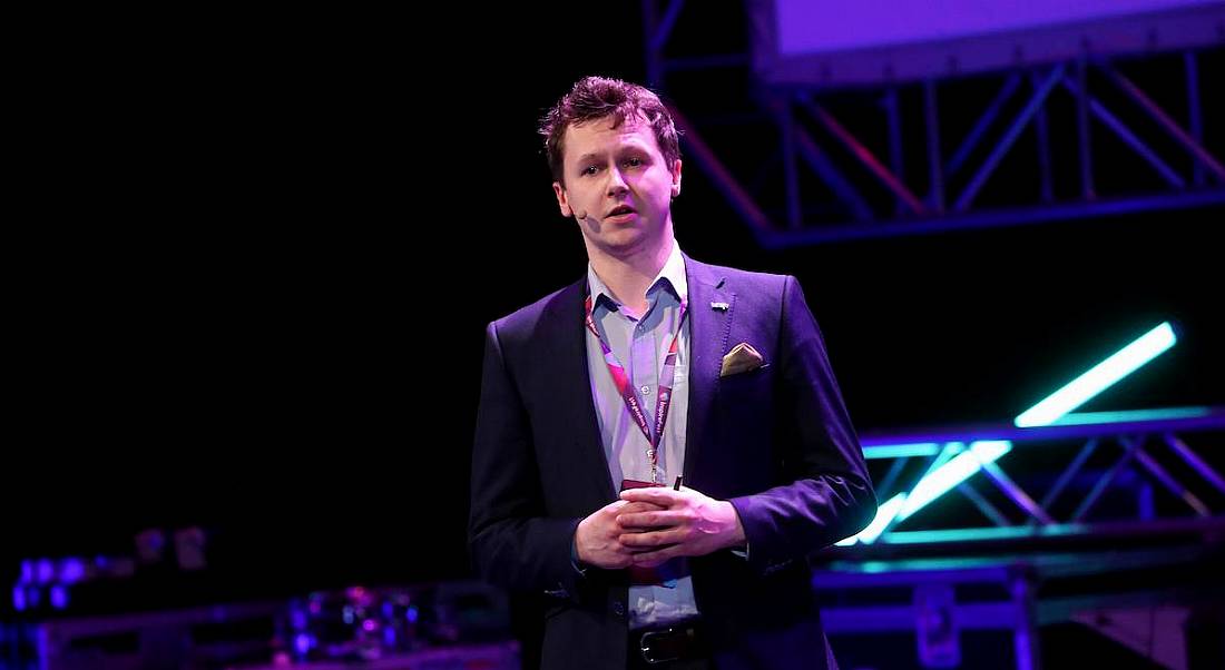 young man wearing pale blue shirt and navy suit speaking on stage into head mic.