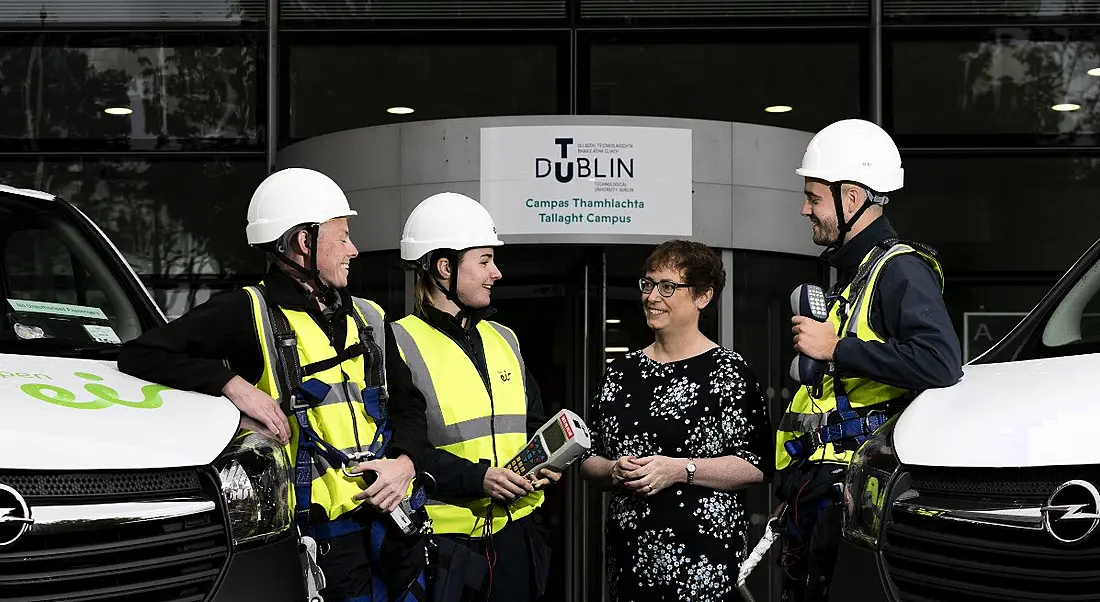 Group of Eir apprentices in hard hats and high-vis gear laughing and chatting with a woman with brown cropped hair and glasses.