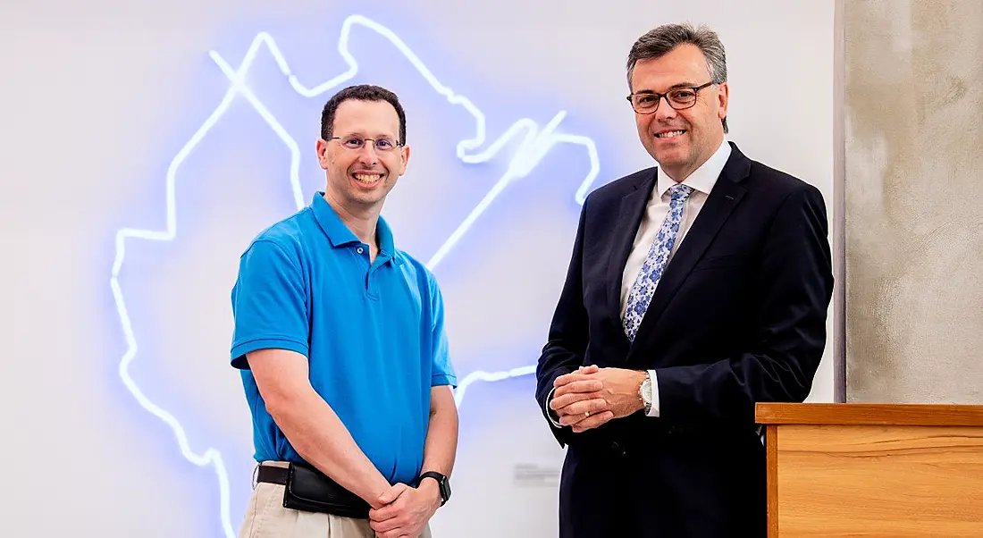 Two men, one in an electric blue polo shirt and slacks, and the other in a business suit, standing and smiling in front of a neon lights art piece.