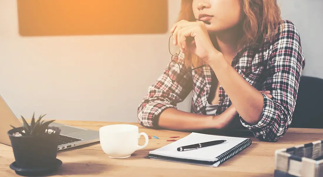 Woman in checked shirt sitting at desk holding her glasses and mulling looking for a new job.