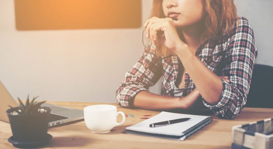 Woman in checked shirt sitting at desk holding her glasses and mulling looking for a new job.