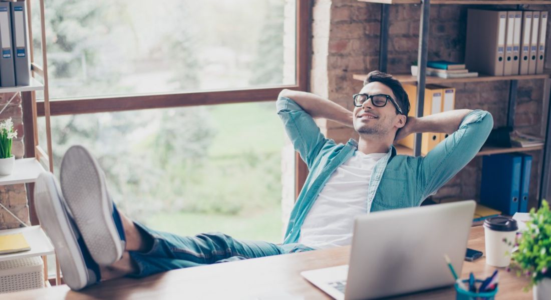 Young man at work wearing shirt and horn-rimmed thick-framed glasses relaxing at desk with feet up.