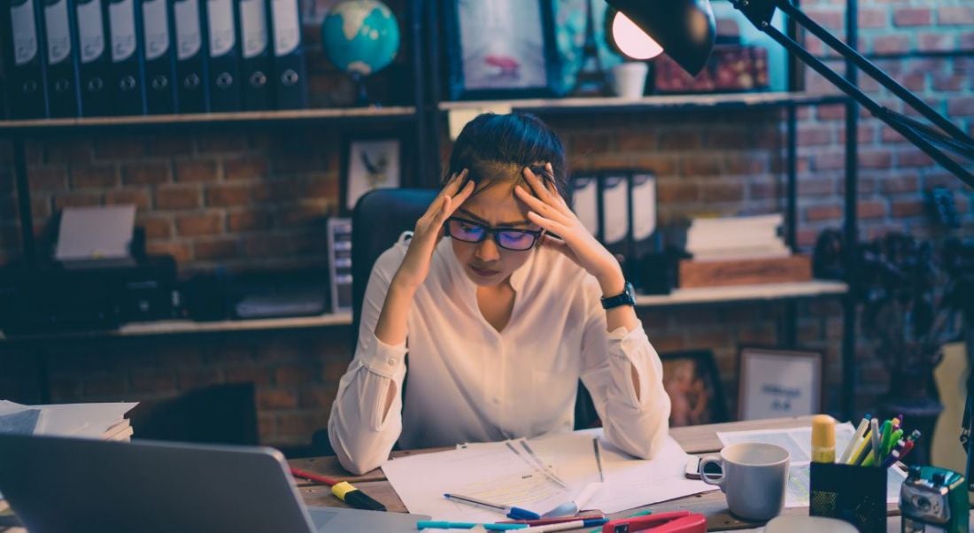 View of young woman in glasses with her hands on her head and brow furrowed with stress, poring over work.