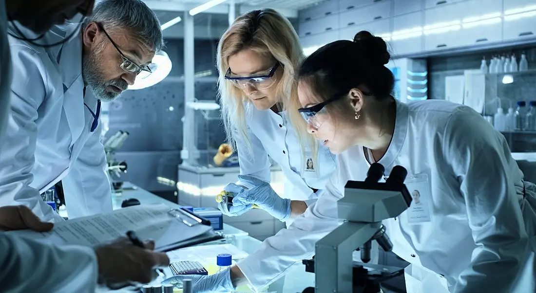 Three scientists, one man and two women, in a laboratory with microscopes and other research equipment.