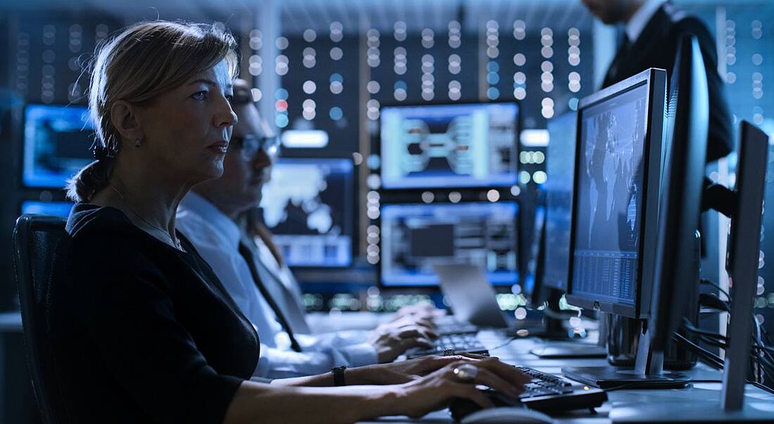 A cybersecurity professional in dark room typing away on her keyboard looking at dual screen computer set up.