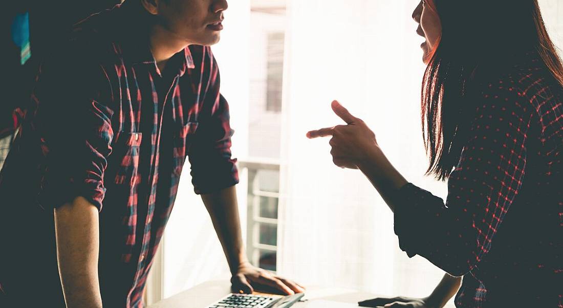 Two people in checked shirts leaning over desk with calculators and papers having an argument in professional setting.