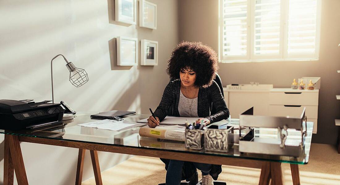 Woman sitting at desk in living room with plush carpet doing work and holding a pen.