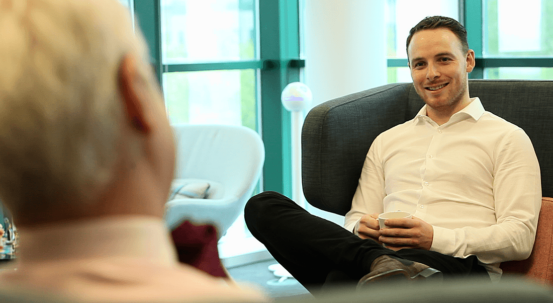 Young man with slicked back brunette hair in crisp white shirt sitting on couch and smiling.