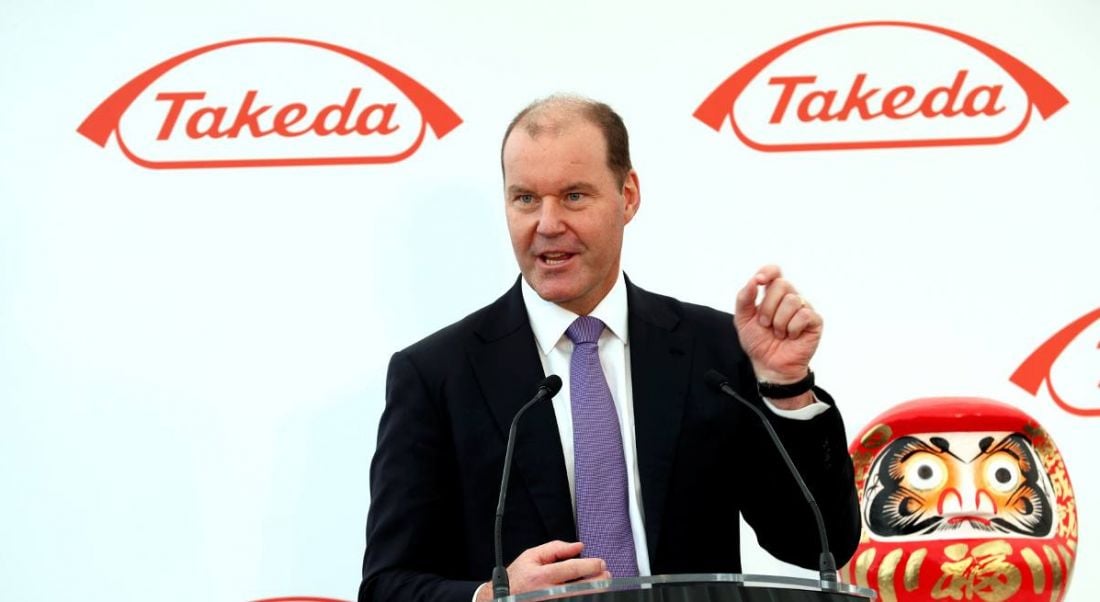 View of man in business suit standing at podium giving speech against backdrop emblazoned with Takeda logo.