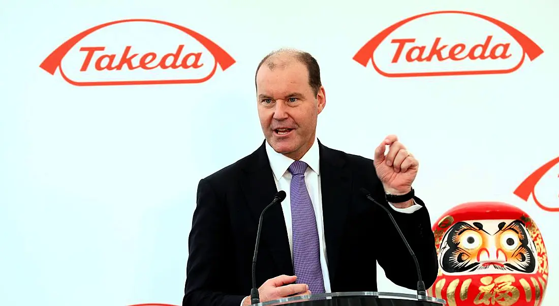 View of man in business suit standing at podium giving speech against backdrop emblazoned with Takeda logo.