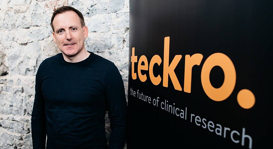 Young man in navy sweater standing next to sign with Teckro logo.