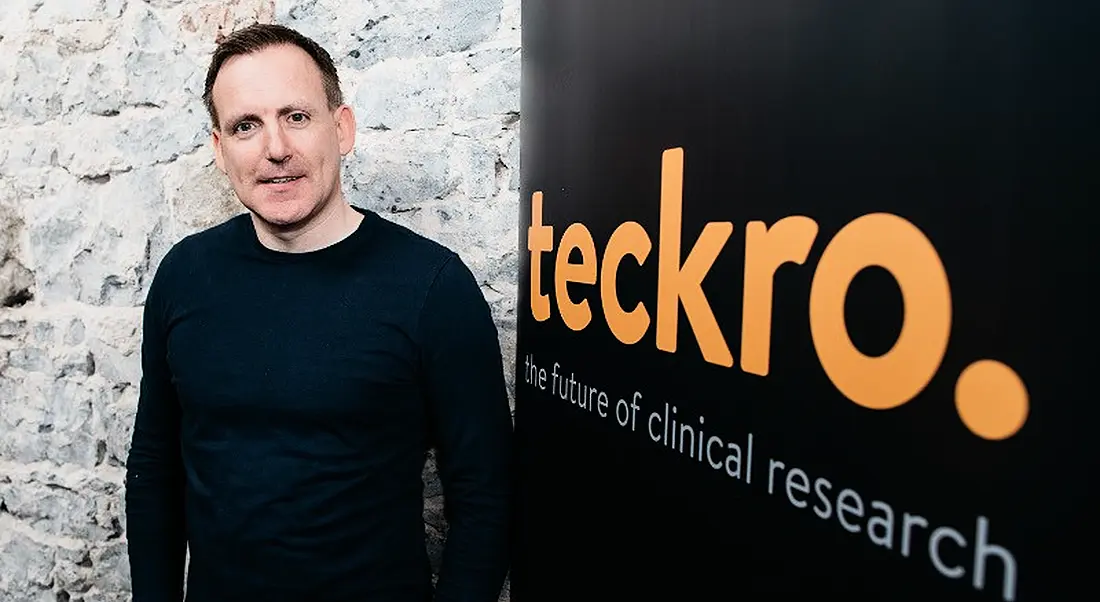 Young man in navy sweater standing next to sign with Teckro logo.