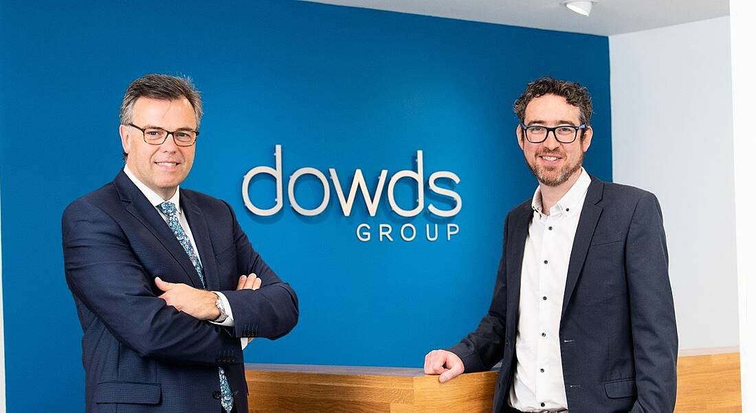 View of two men in business suits, one slate grey and one navy, standing in front of a blue wall with Dowds Group logo on it.