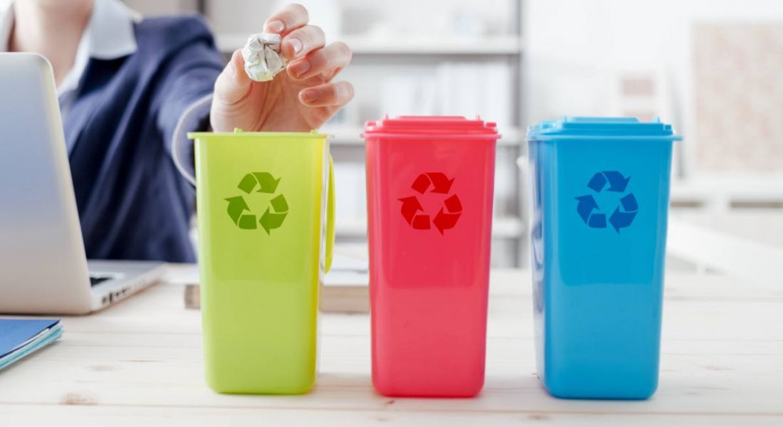 close-up of woman's hand putting paper waste into a mini green bin on her office desk. There is also pink bin and blue bin, all denoting the recycling logo.