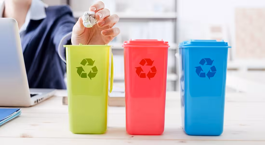 close-up of woman's hand putting paper waste into a mini green bin on her office desk. There is also pink bin and blue bin, all denoting the recycling logo.