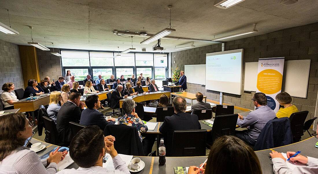 group of people sitting in a meeting room watching man in suit give a talk.