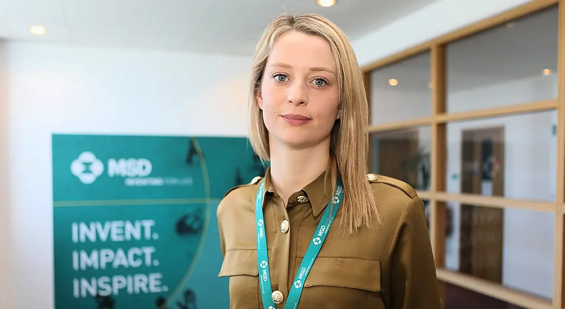 Young blonde woman with brown shirt wearing turquoise lanyard standing in front of MSD banner.