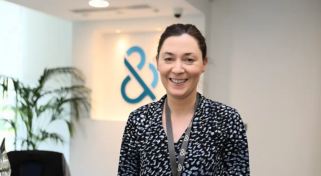 Woman in polka dot navy blouse and lanyard standing in modern office and smiling.