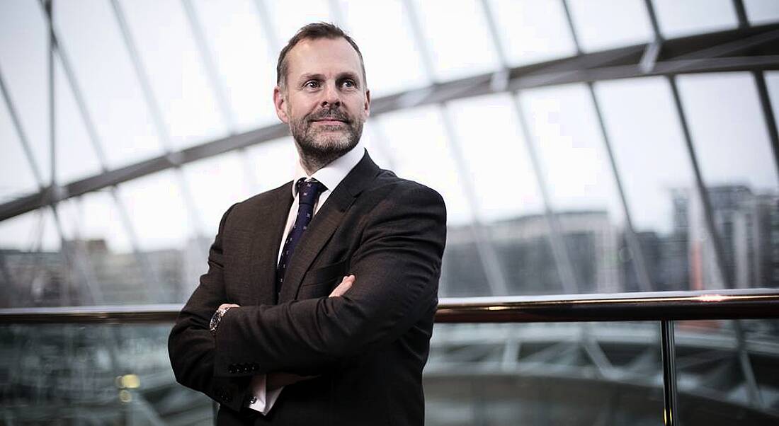 A man standing in a business suit with arms folded looking beyond the camera in front of large windows in a corporate space.