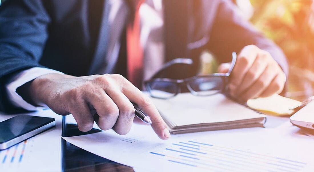 View of hands, one holding thick-rimmed glasses and the other floating over professional documents, at a dark wood desk.