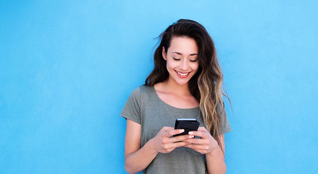 View of young woman with tousled ombré hair and grey shirt holding phone in hands and smiling down at it.