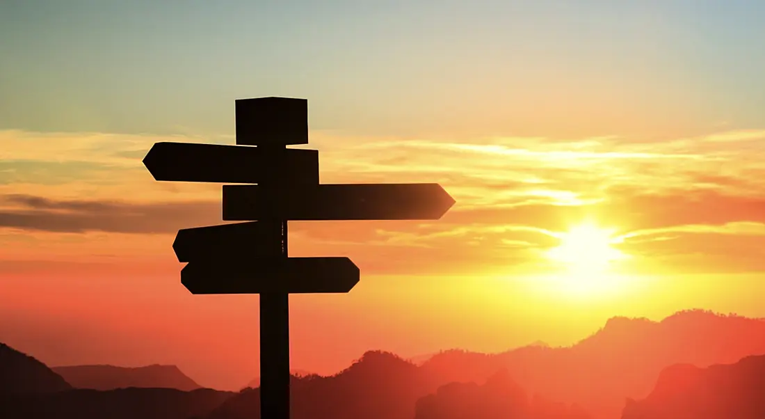 View of sign post indicating different directions, set against colourful sunset.