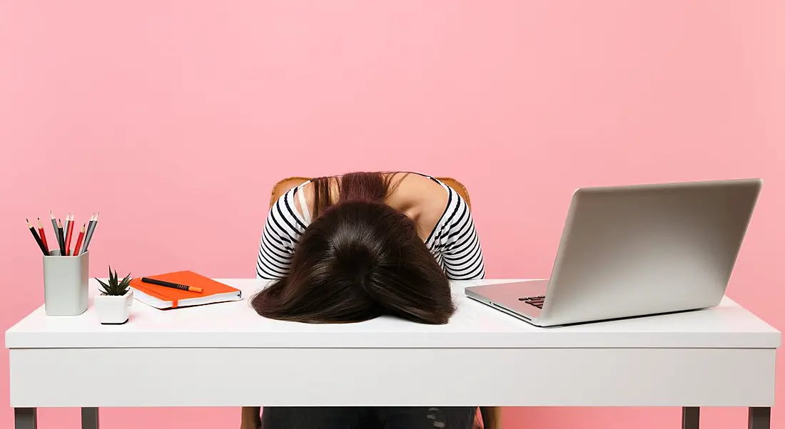 View of woman at work desk with head on the table experiencing burnout against bubblegum pink background.