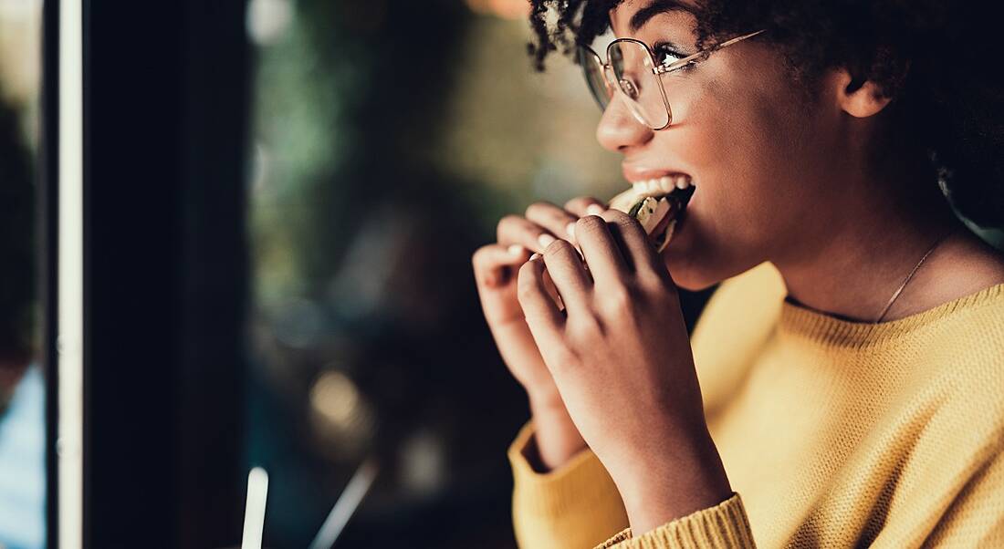 View of young woman with glasses smiling and biting into a sandwich on her lunch break.