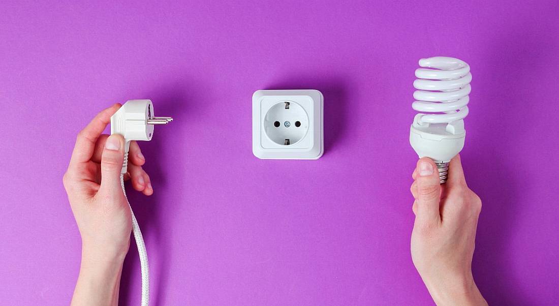 View of hands, one holding a light bulb and the other holding a cord, with a European plug in the middle against a vibrant violet background.