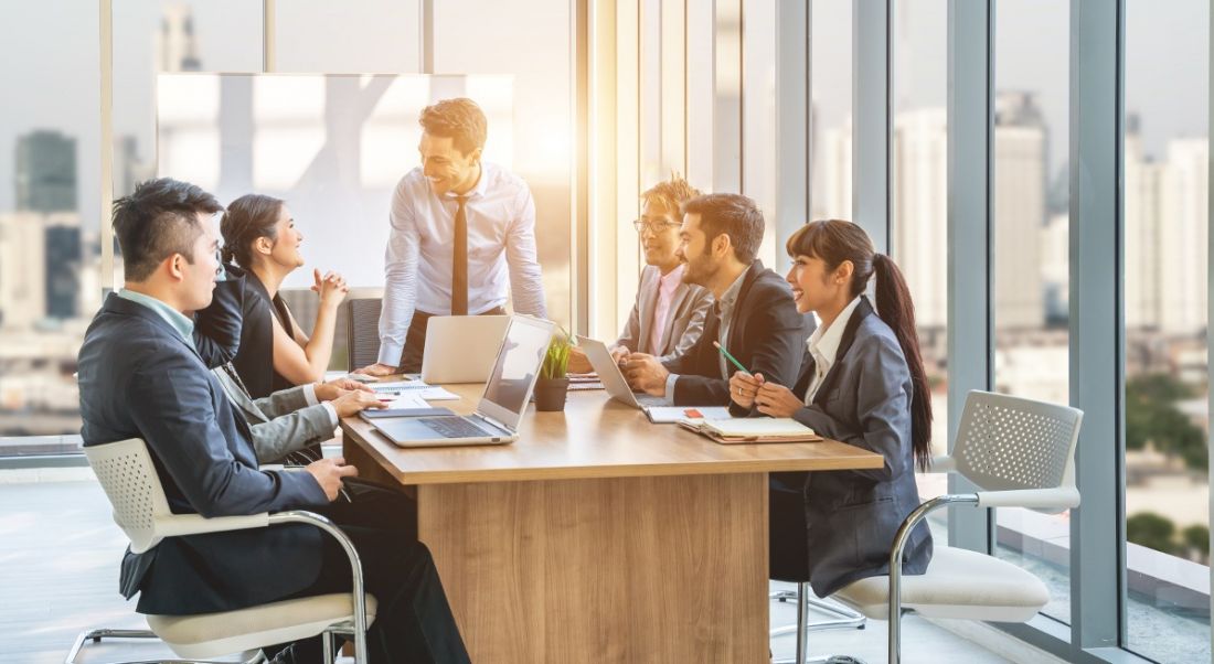 people in business attire in room with high ceilings smiling gathered around a table having a meeting.