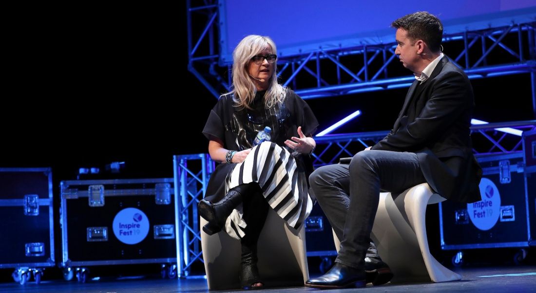 A man and woman are sitting close to each other and talking on a stage with purple lighting behind them.