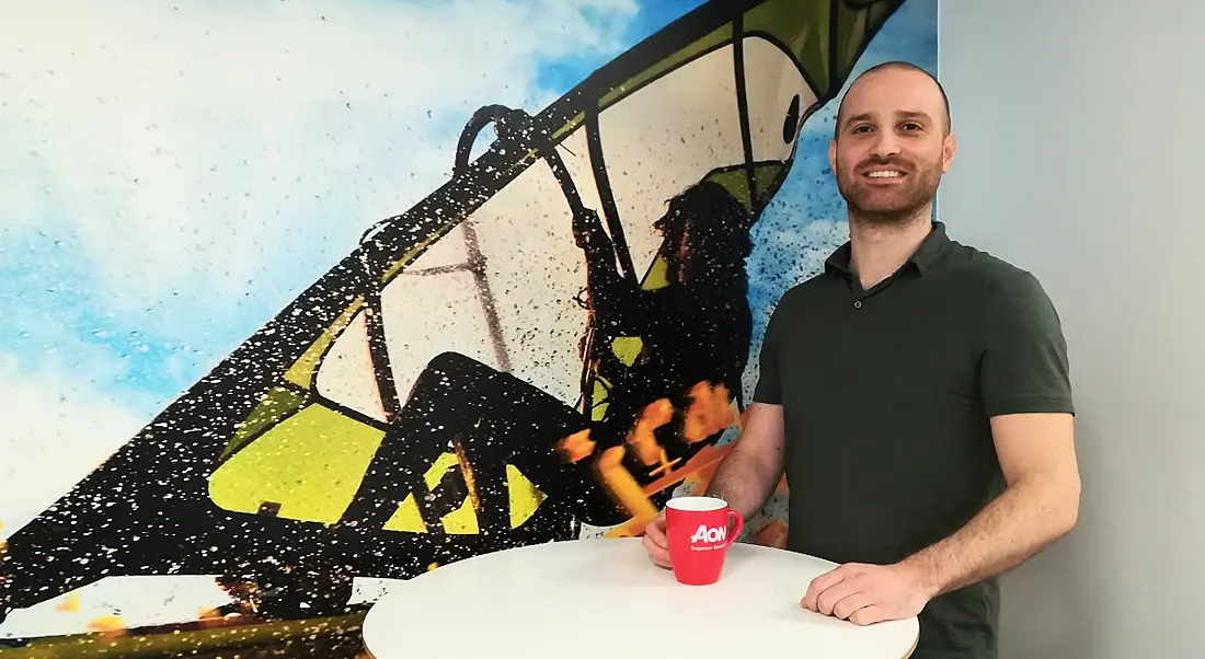 Young man in green T-shirt holding Aon mug and smiling against graphic mural background.