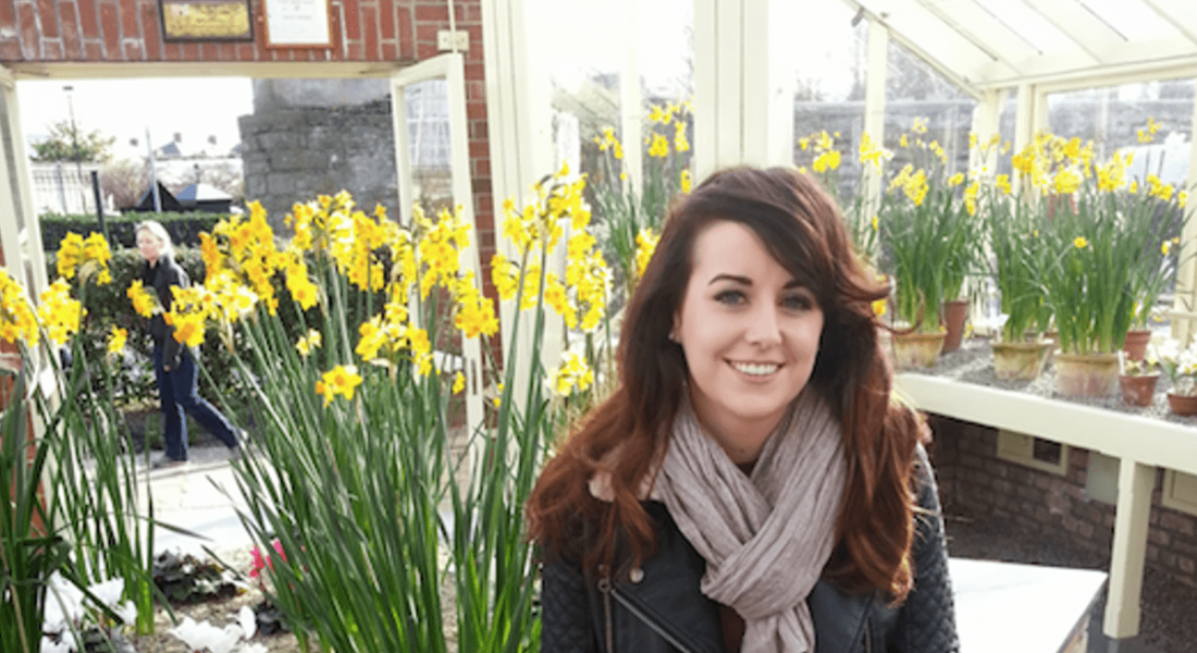 View of brunette young woman swathed in a beige scarf sitting against a backdrop of blooming daffodils in a conservatory.