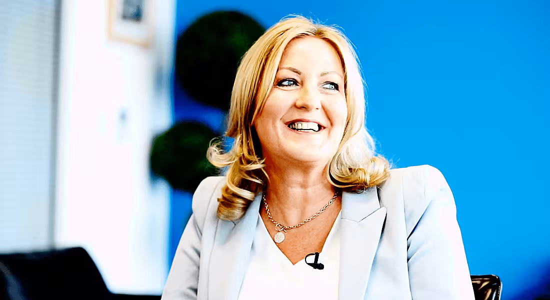 View of smiling blonde woman in light blue pant suit looking off camera in electric blue room.