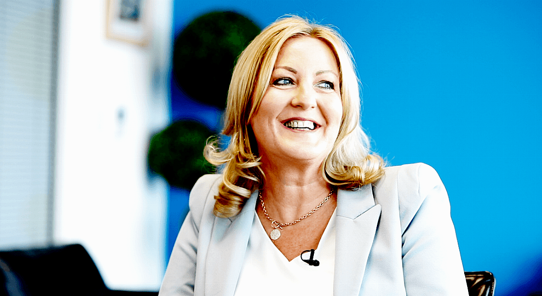 View of smiling blonde woman in light blue pant suit looking off camera in electric blue room.