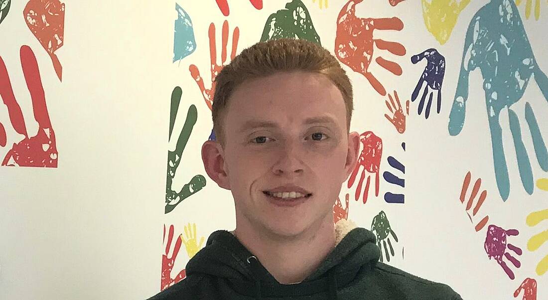 A young man in a green hoodie stands in front of a wall that is covered with hand prints.