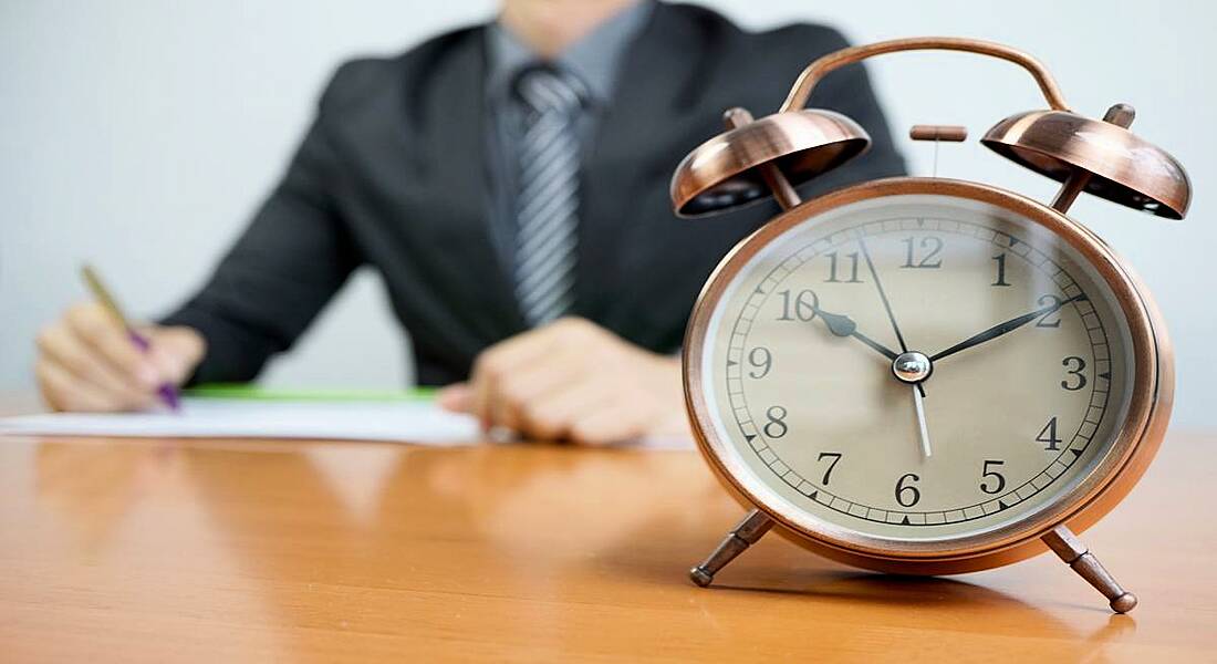 A businessman in a suit sitting at a desk behind an alarm clock with is the focus of the photo.