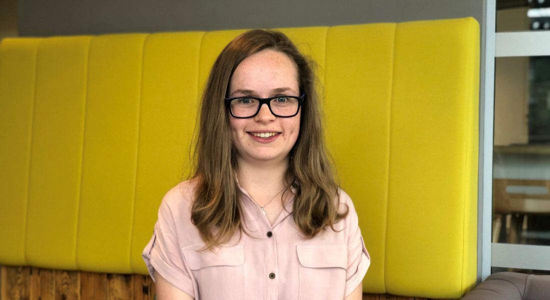 View of young woman in pale pink blouse with glasses smiling at camera against yellow background.