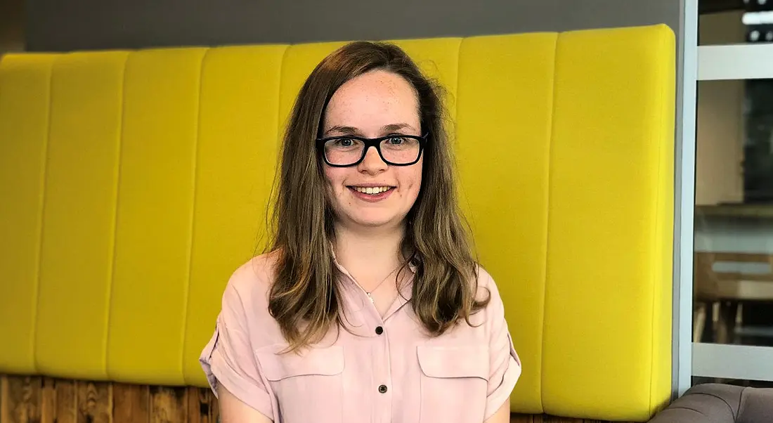 View of young woman in pale pink blouse with glasses smiling at camera against yellow background.