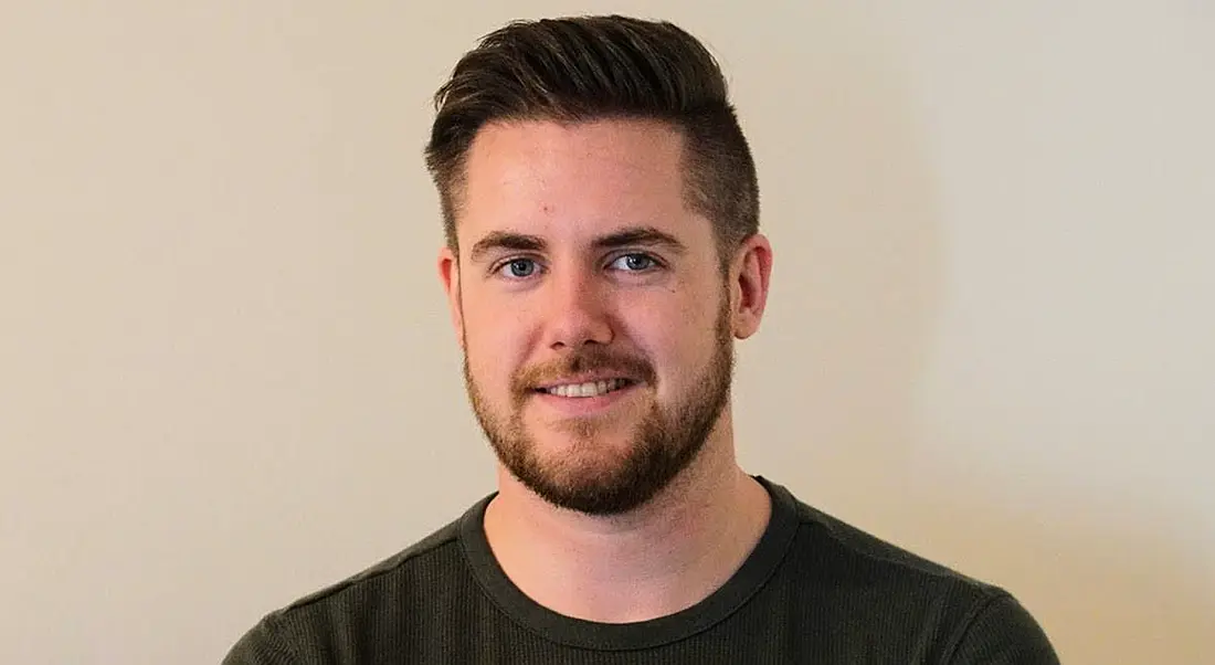 A headshot of a young male wearing a dark green T-shirt and standing against a plain wall.