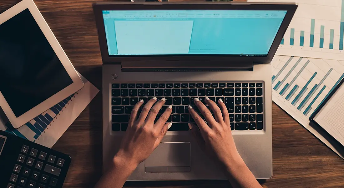 Bird's eye view of female hands typing on a laptop on a desk with notes around it.