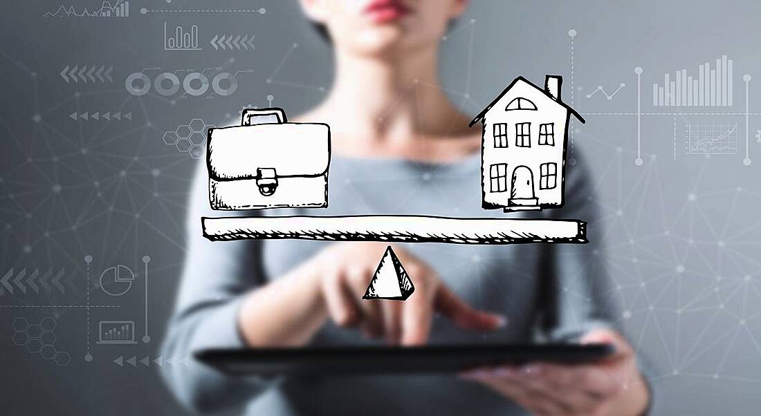 Woman is standing in front of a grey background and behind an illustration of a scale with a briefcase on the left and a house on the right.