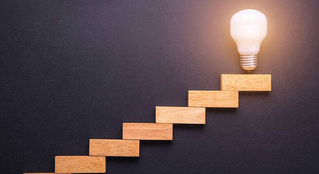 Wooden blocks set up as a staircase with light bulb on the top, in front of black stone board.