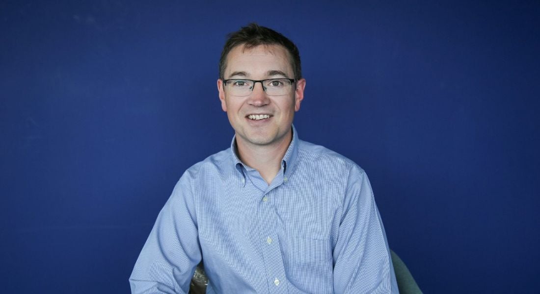 View of man with glasses in blue shirt smiling sitting against cobalt blue wall.