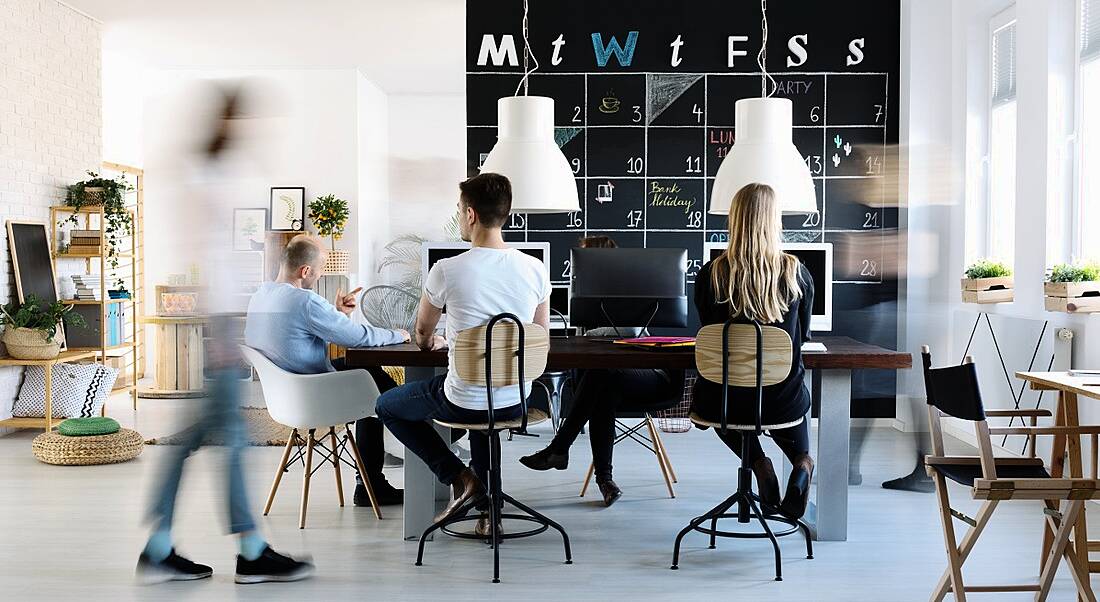 A creative open-plan office space with monthly planner in chalk on a back wall and people milling around desks and computers.